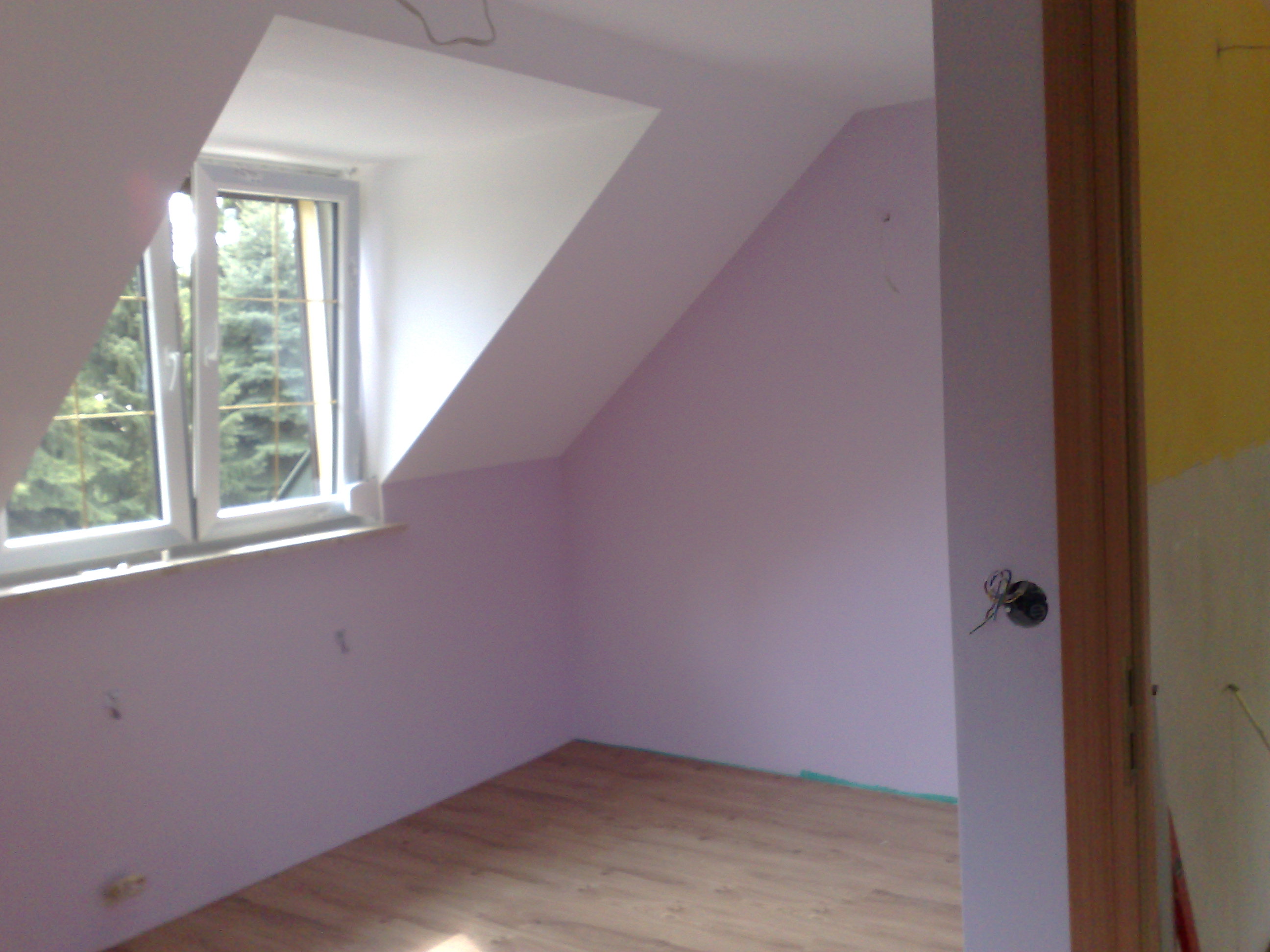 Pokój na poddaszu z wykończonymi ścianami w odcieniu liliowym, nową drewnianą podłogą i oknem dachowym z widokiem na drzewa, widoczne przewody elektryczne przy drzwiach.